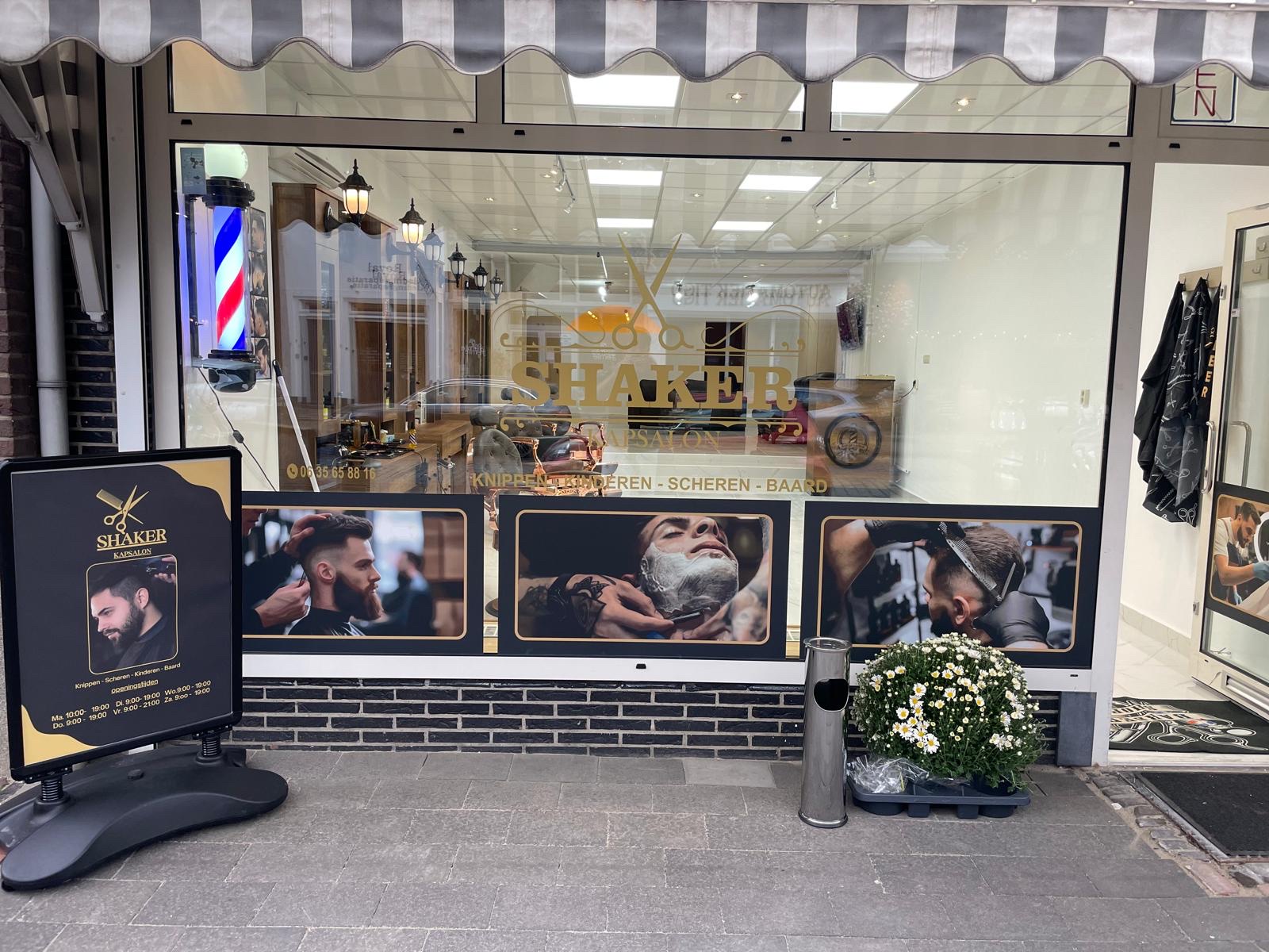 De etalage van Shaker Kapsalon aan de Waterstraat, met de gouden belettering, de barbierspaal en het stoepbord